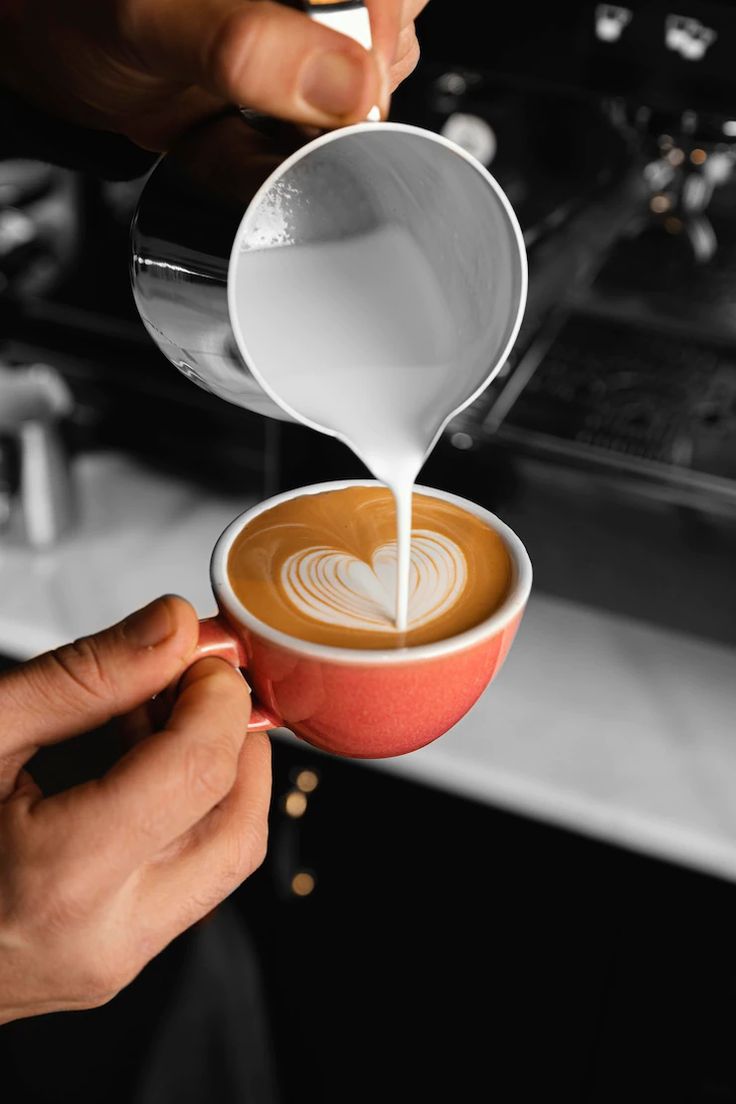 Hands pouring steamed milk from a metal pitcher into a coral-pink cup with heart-shaped latte art